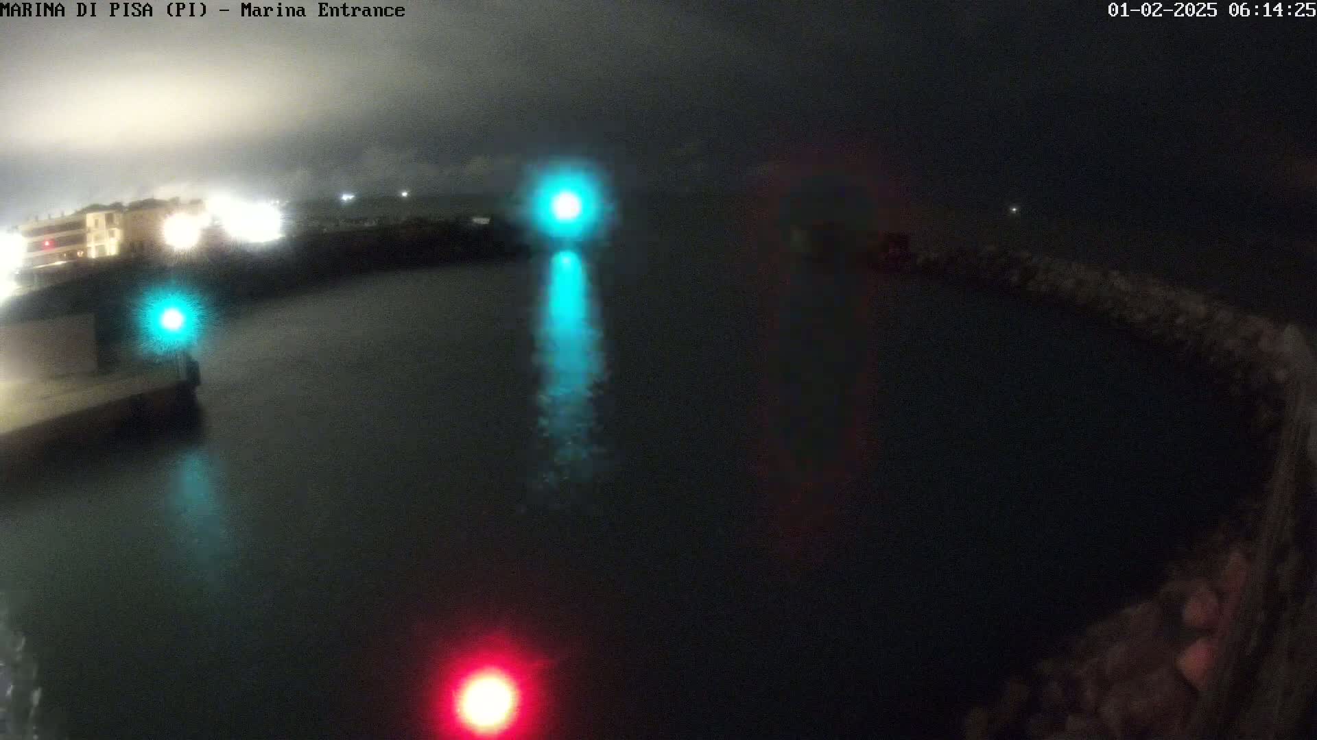 A nighttime, dark view of a harbor entrance with a teal and a red light visible on the water, under a cloudy sky.