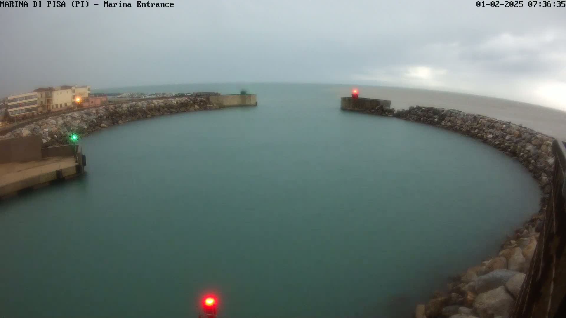 A calm, teal-colored harbor is enclosed by stone walls and marked by red and green lights under an overcast sky.