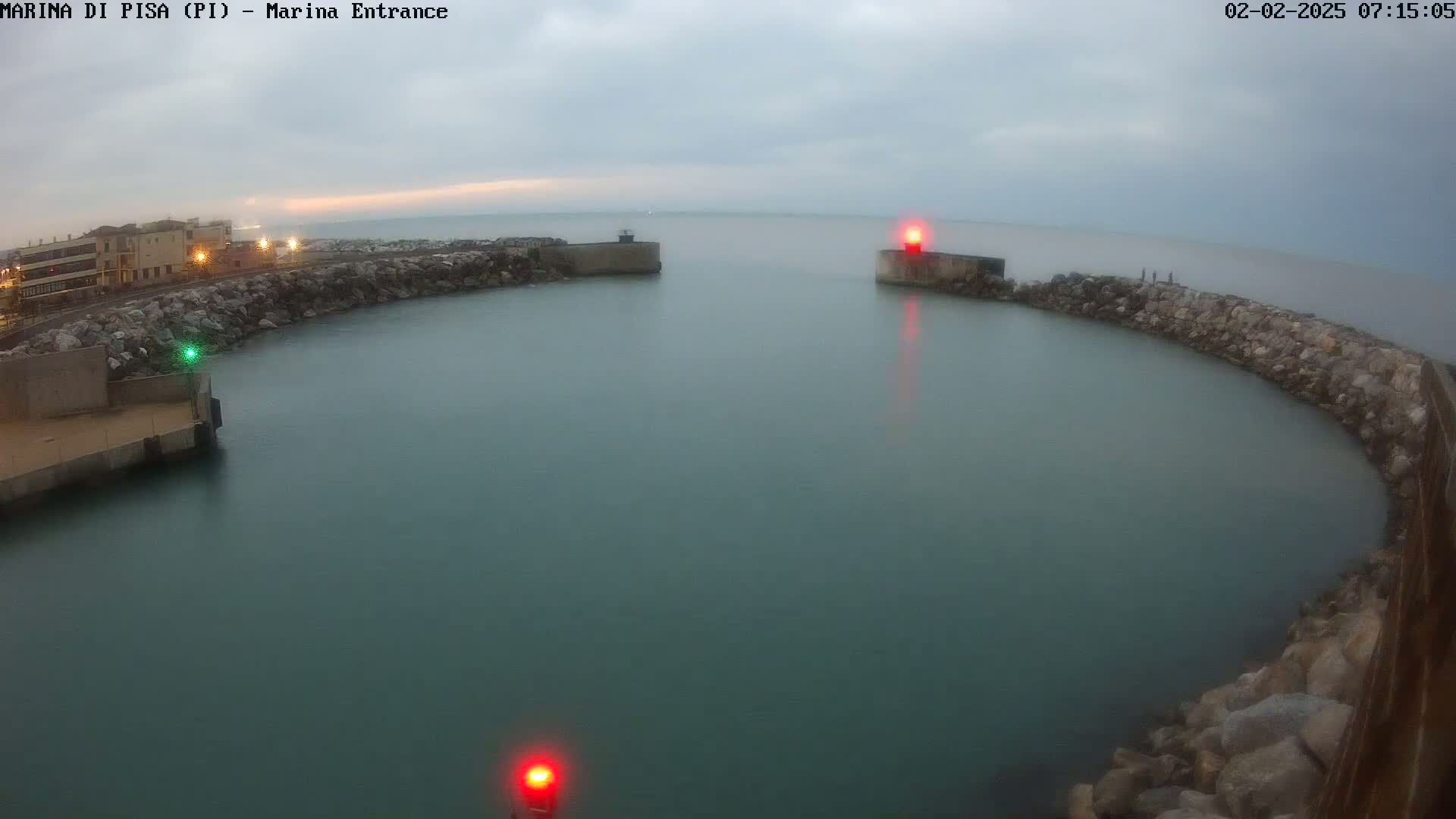 A calm, overcast day reveals a harbor entrance with a curved stone wall,  two red navigation lights, and a building visible in the distance.