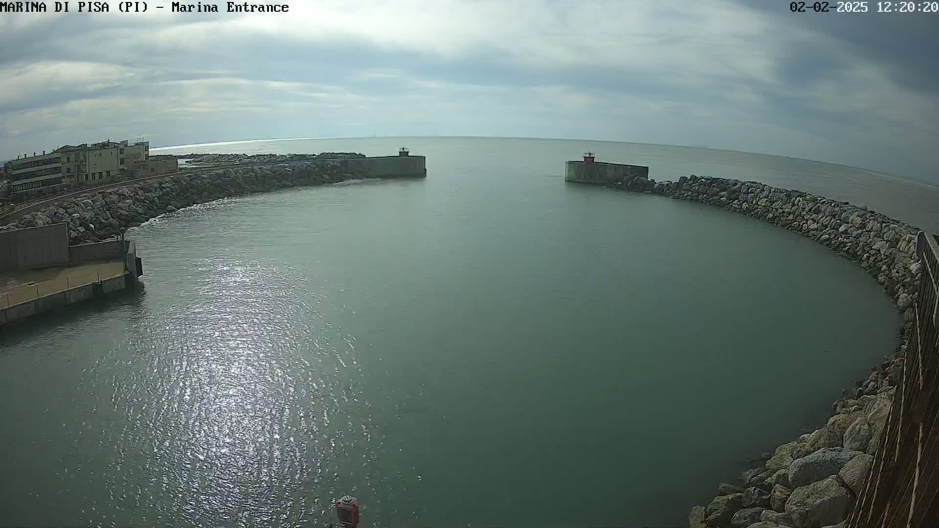 A partially cloudy day shows a harbor entrance with calm, greenish water, bordered by rocky walls and buildings.