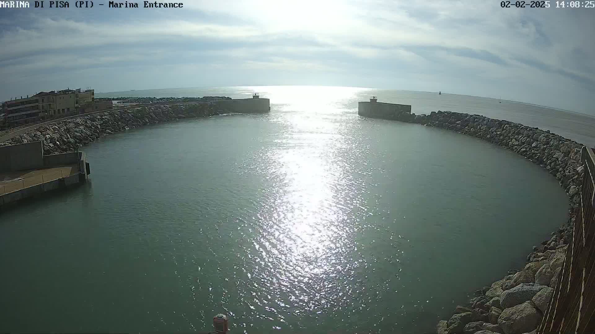 A sunny day reveals a harbor entrance with calm, sun-lit water enclosed by rock walls and a building in the background.