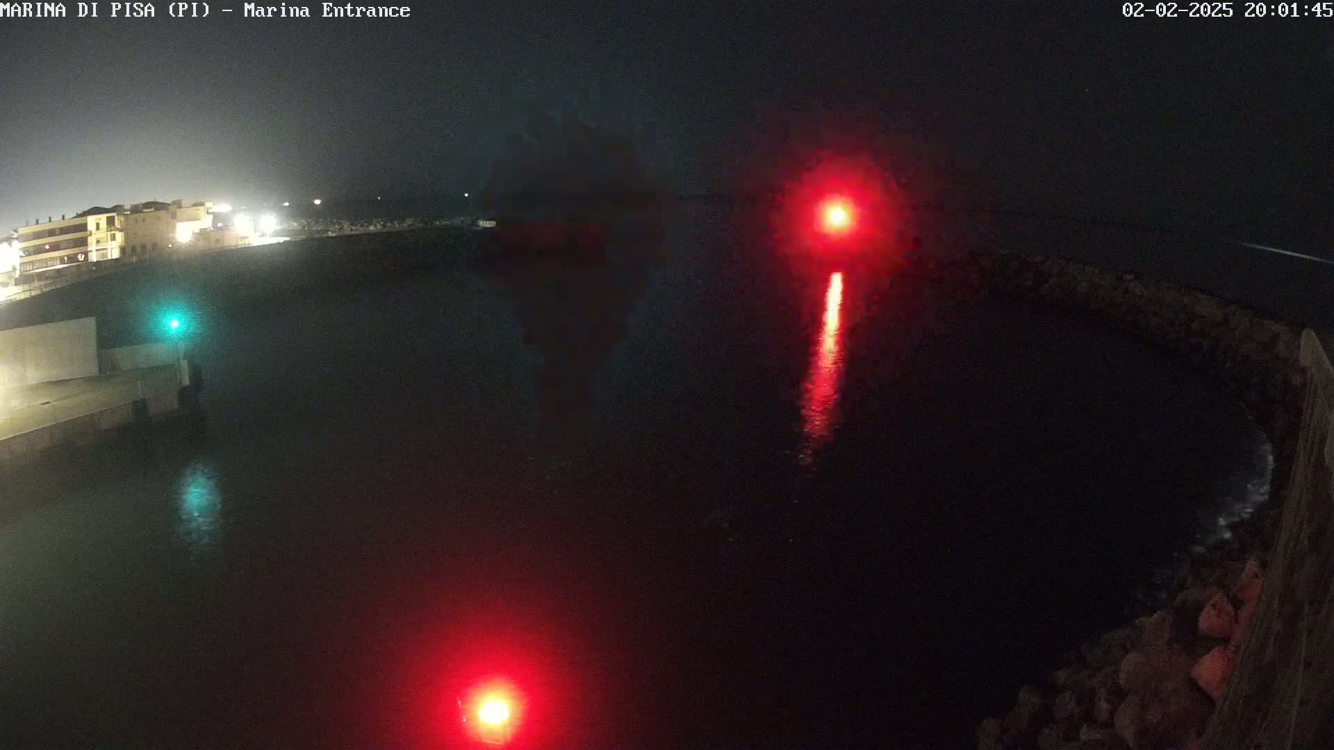A nighttime view of a dark body of water with red and green lights reflecting on the surface, illuminated buildings in the background, and a stone wall on the right.