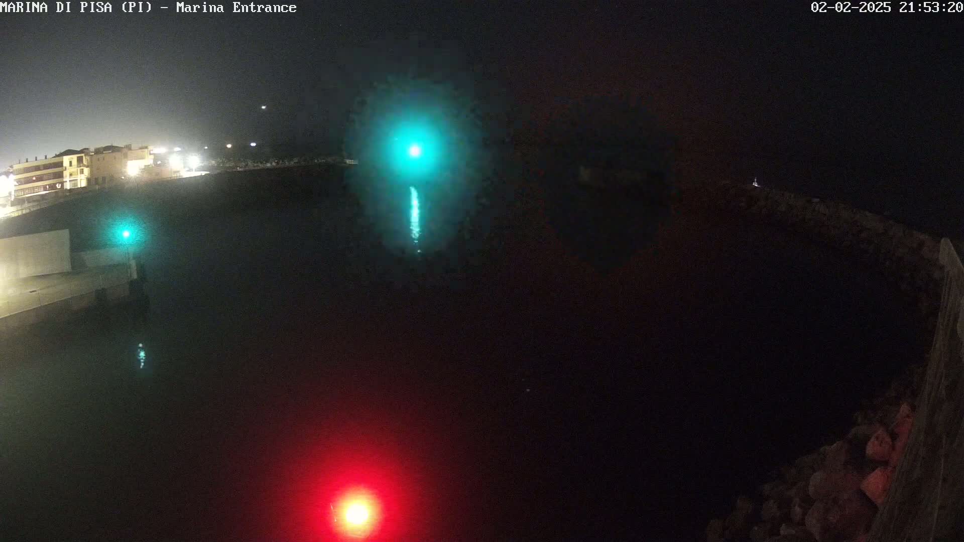 A nighttime, high-angle view shows a dark body of water with a green and a red light visible near the edges, and a building is visible in the distance.