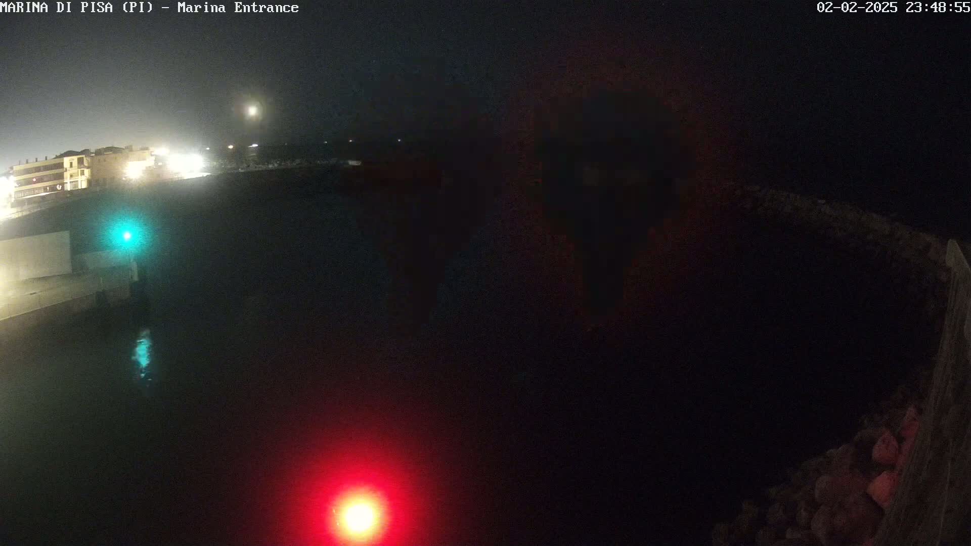 A nighttime view of a dark body of water with faintly lit buildings in the background and a red and green light visible near the water's edge.