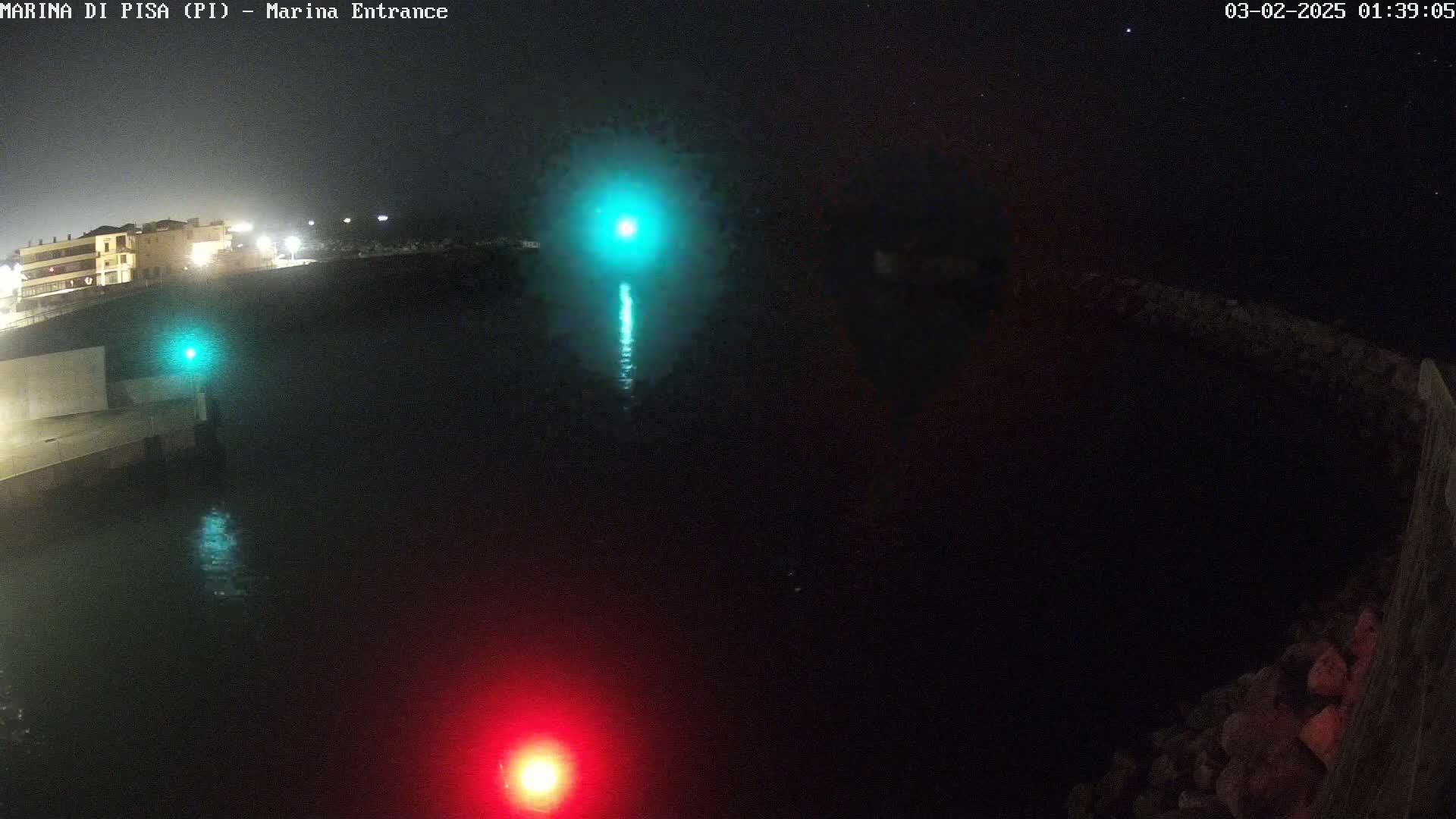 A nighttime aerial view of a marina entrance shows dark water reflecting a green and a red light, with a building visible in the distance under a clear, starry sky.
