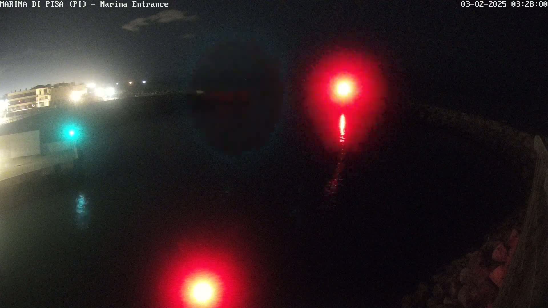 At night, two red lights and one green light illuminate a dark body of water near a shoreline with buildings in the distance under a mostly clear sky.