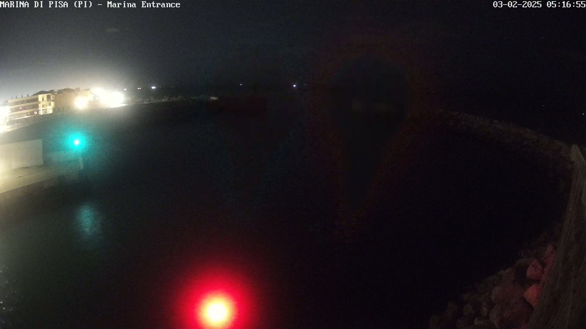 A nighttime, dark view of a marina entrance with a red and green light visible on the water, and faintly lit buildings in the distance.