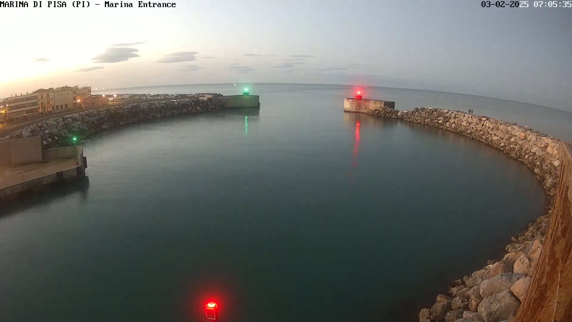 A calm, dark-blue body of water is surrounded by a curved rock wall, with a building visible on the left and two light posts marking a channel entrance under a dusky sky.