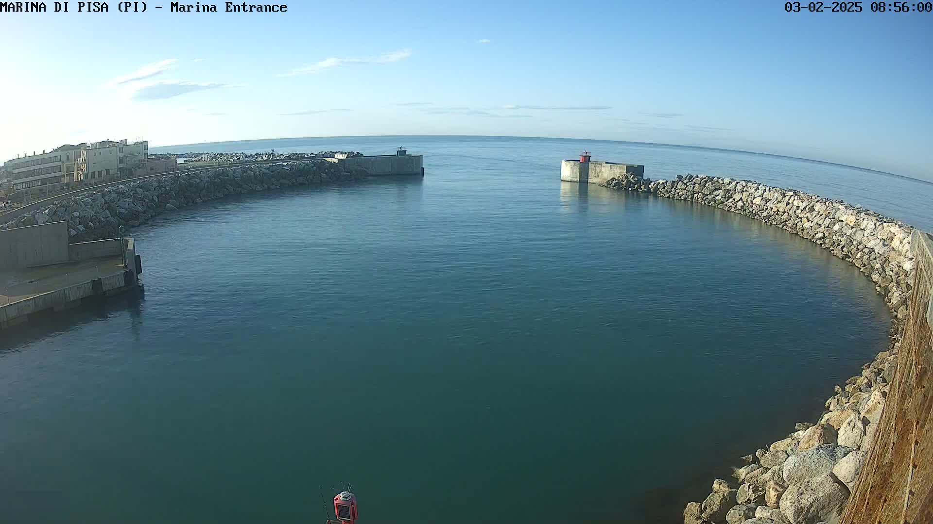 A calm, clear day reveals a harbor entrance with stone breakwaters, a small building in the distance, and a buoy in the water.