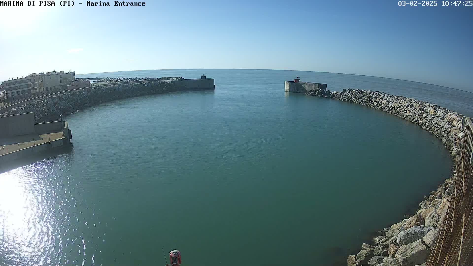 Under a clear blue sky, a calm body of teal water is enclosed by two stone breakwaters, with a building visible on one side.