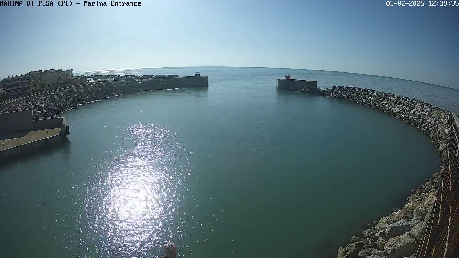 A sunny day reveals a calm harbor entrance, bordered by rocky breakwaters and a building in the distance.