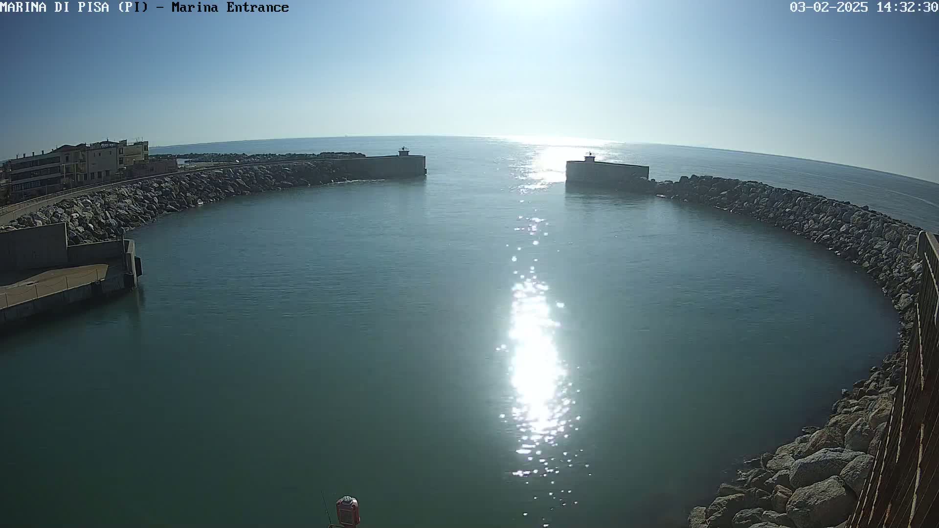 A sunny day reveals a calm harbor entrance defined by rock walls, with the sun's reflection shimmering on the water's surface.