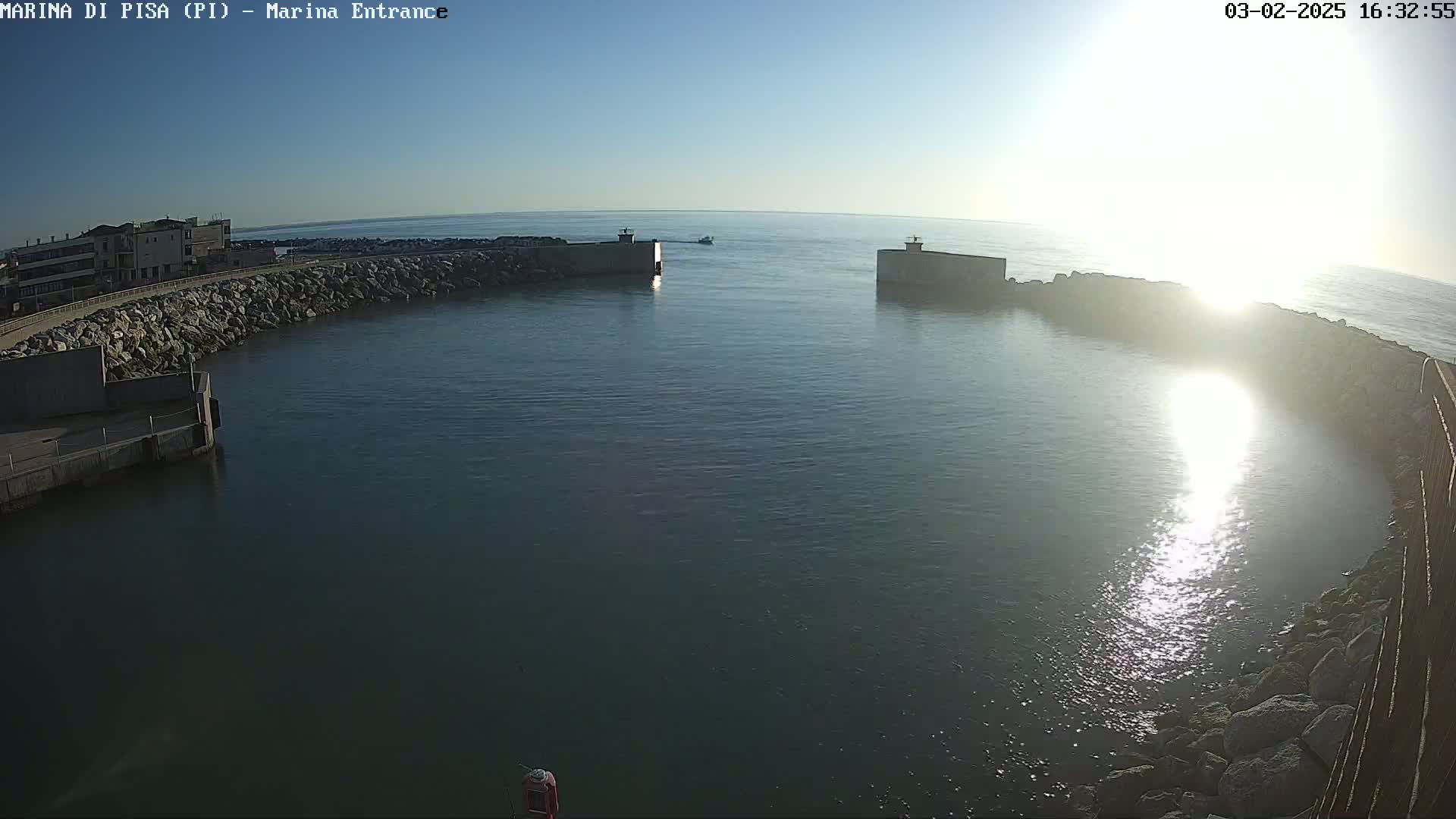A harbor entrance with calm water under a bright, sunny sky is bordered by rocky walls and buildings.