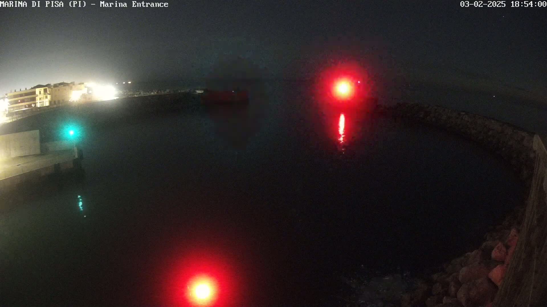 A nighttime harbor scene shows dark water with two red navigation lights and one green light, illuminated buildings in the distance, and a stone breakwater.
