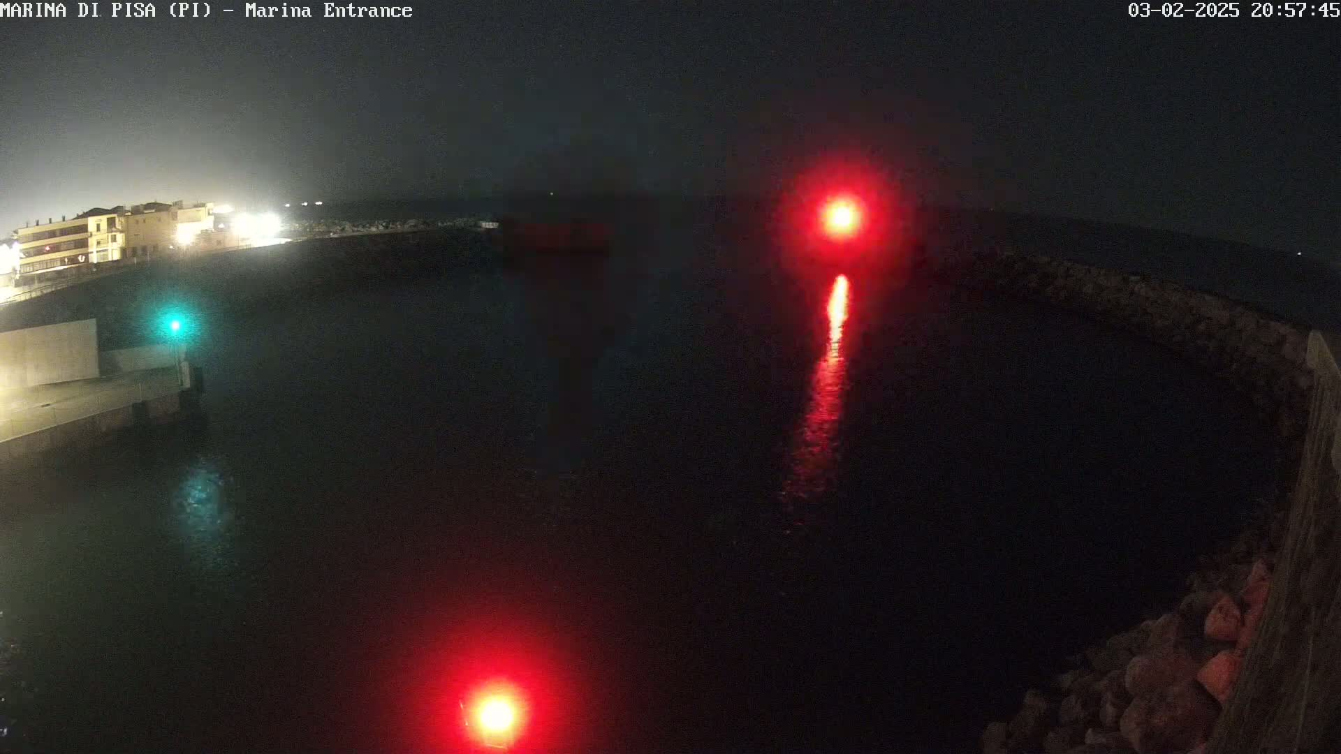 At night, a dark body of water is illuminated by two red lights and a green light near a stone structure and buildings.
