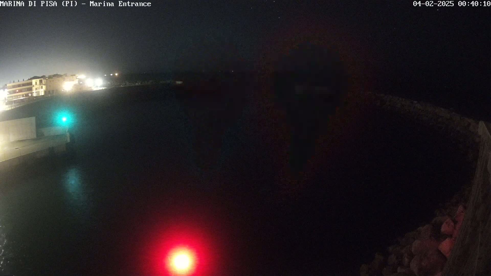 A nighttime, dark view of a harbor entrance with a few lights visible on the buildings and a red and green light on the water.