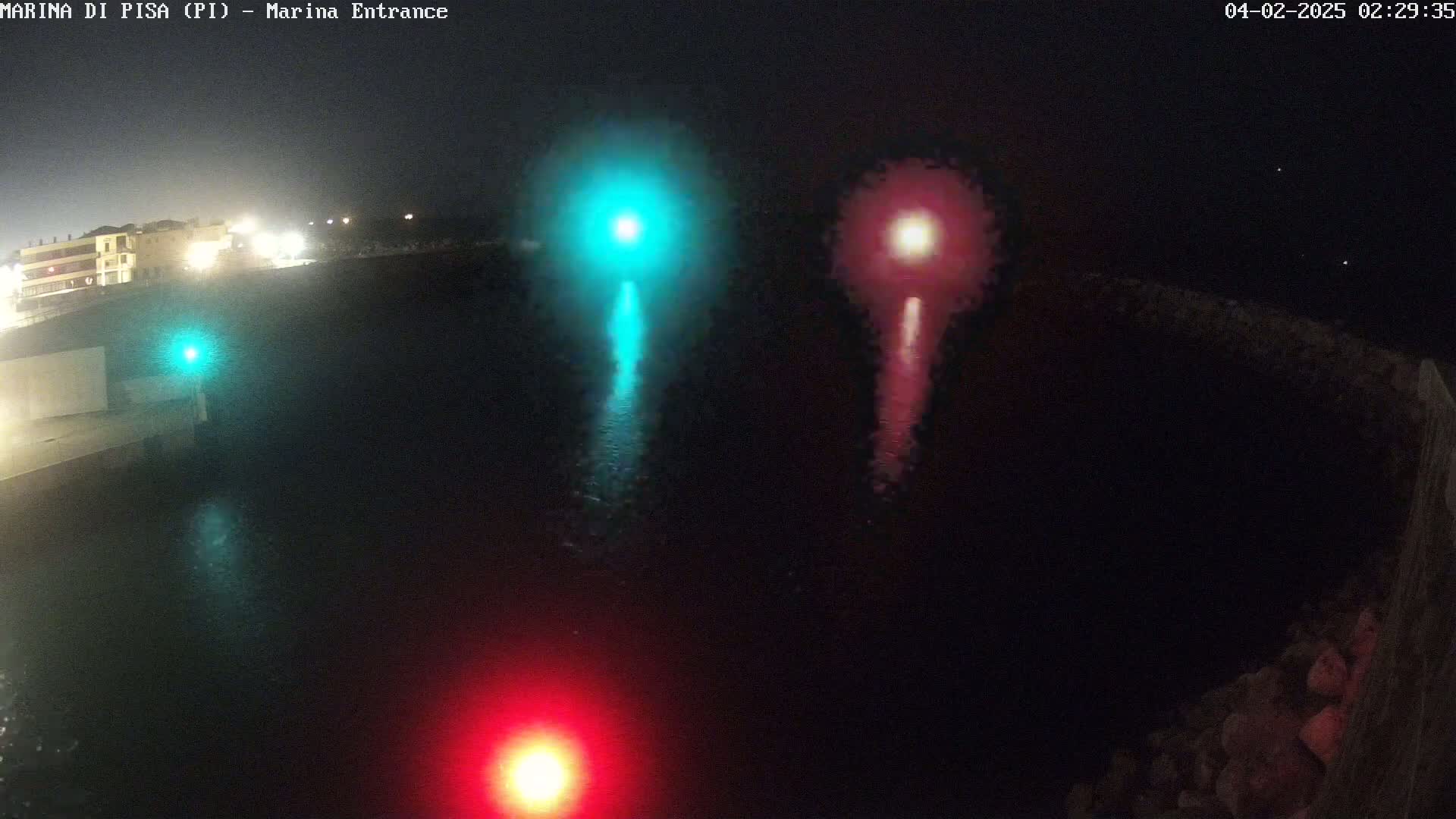 At night, a teal and a red light shine on dark water near a dark stone structure, with a faint glow of city lights in the distance.