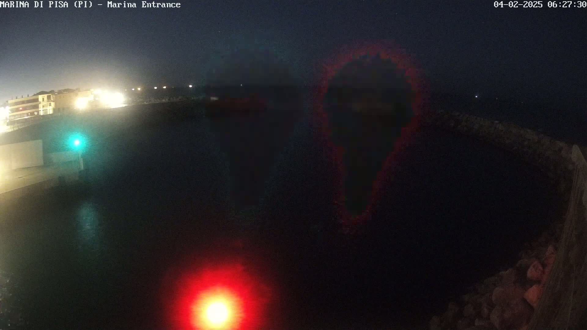 A nighttime, low-light view of a dark body of water, bordered by a stone wall, with a red and green light visible near the shore and faint light sources visible in the distance.