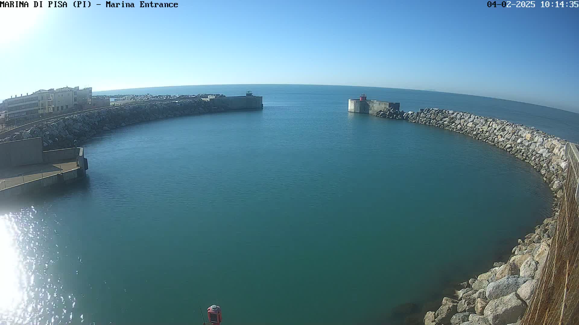 A harbor entrance with calm, teal water is surrounded by rock walls and a building on the left, under a clear blue sky.