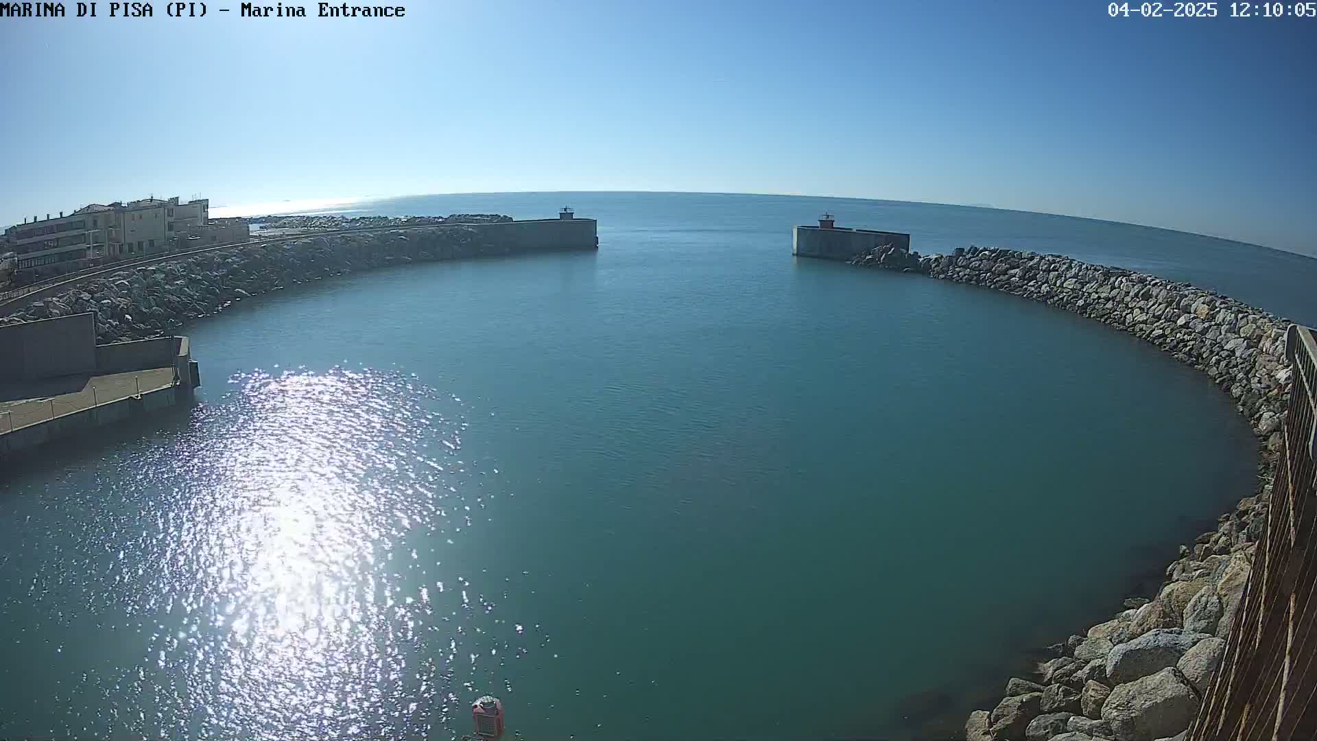 Under a clear blue sky, a calm teal body of water is partially enclosed by a curved rock wall with two concrete structures at its entrance.
