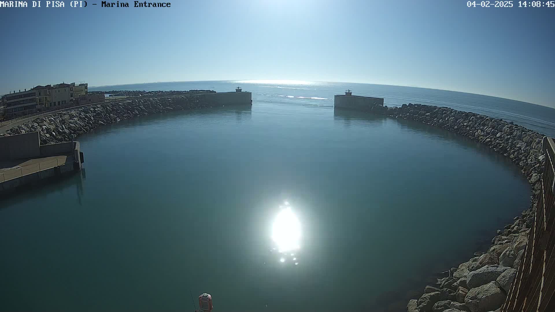 A calm, sunny day shows a harbor entrance with teal water enclosed by rock walls, and a building visible in the distance.