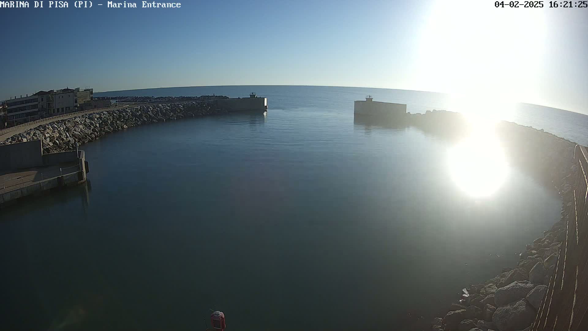 A harbor entrance with calm water is seen on a sunny day, bordered by rocky walls and buildings.