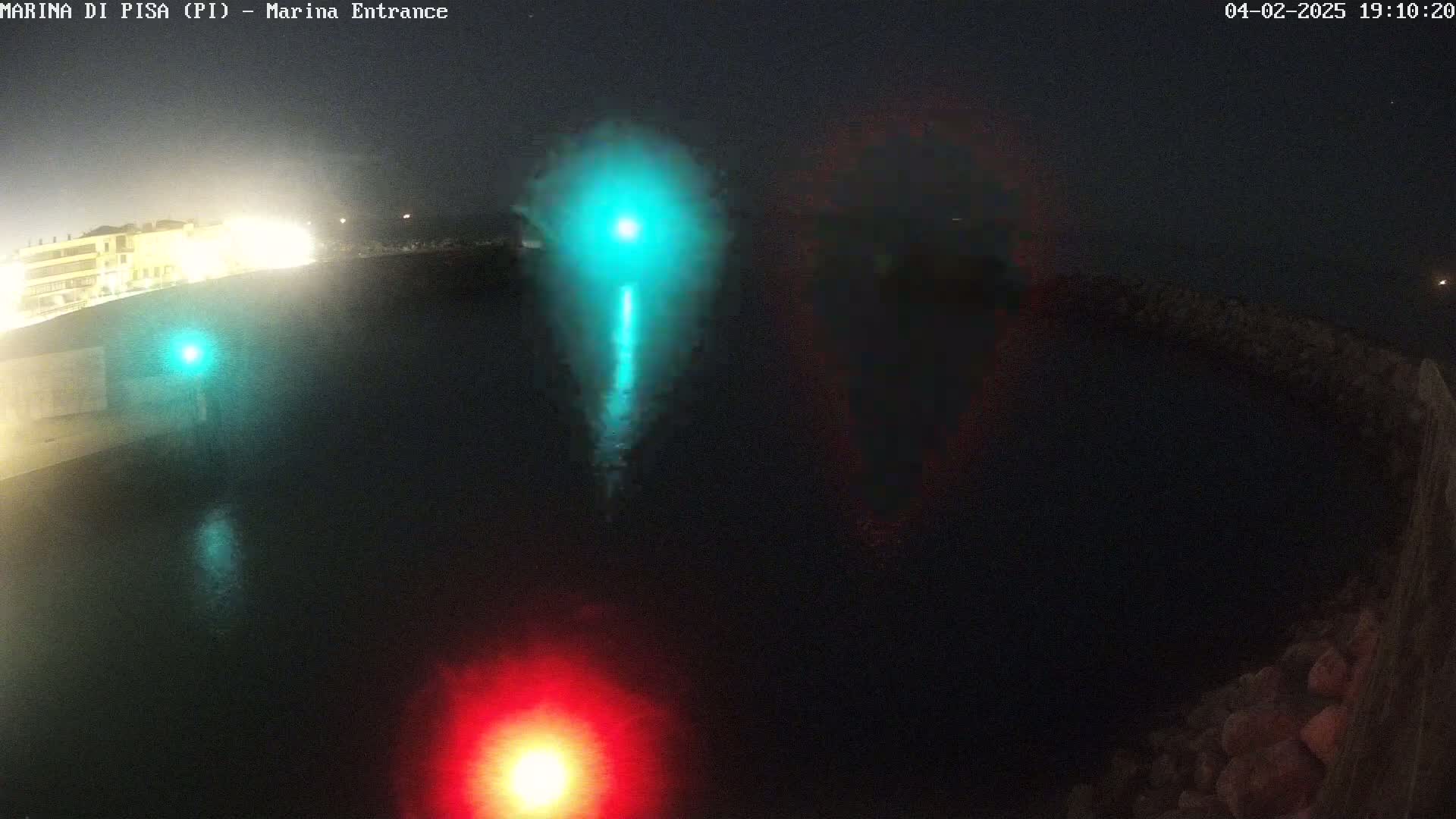 A nighttime view of a dark body of water with a teal and a red light visible on the surface, and dimly lit buildings in the background.