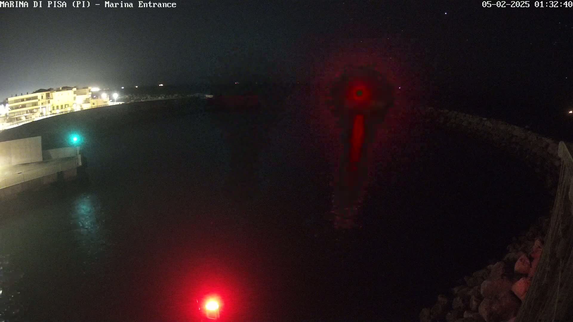 A nighttime, dark view of a marina entrance with red and green navigation lights and a building illuminated in the distance under a clear, starry sky.
