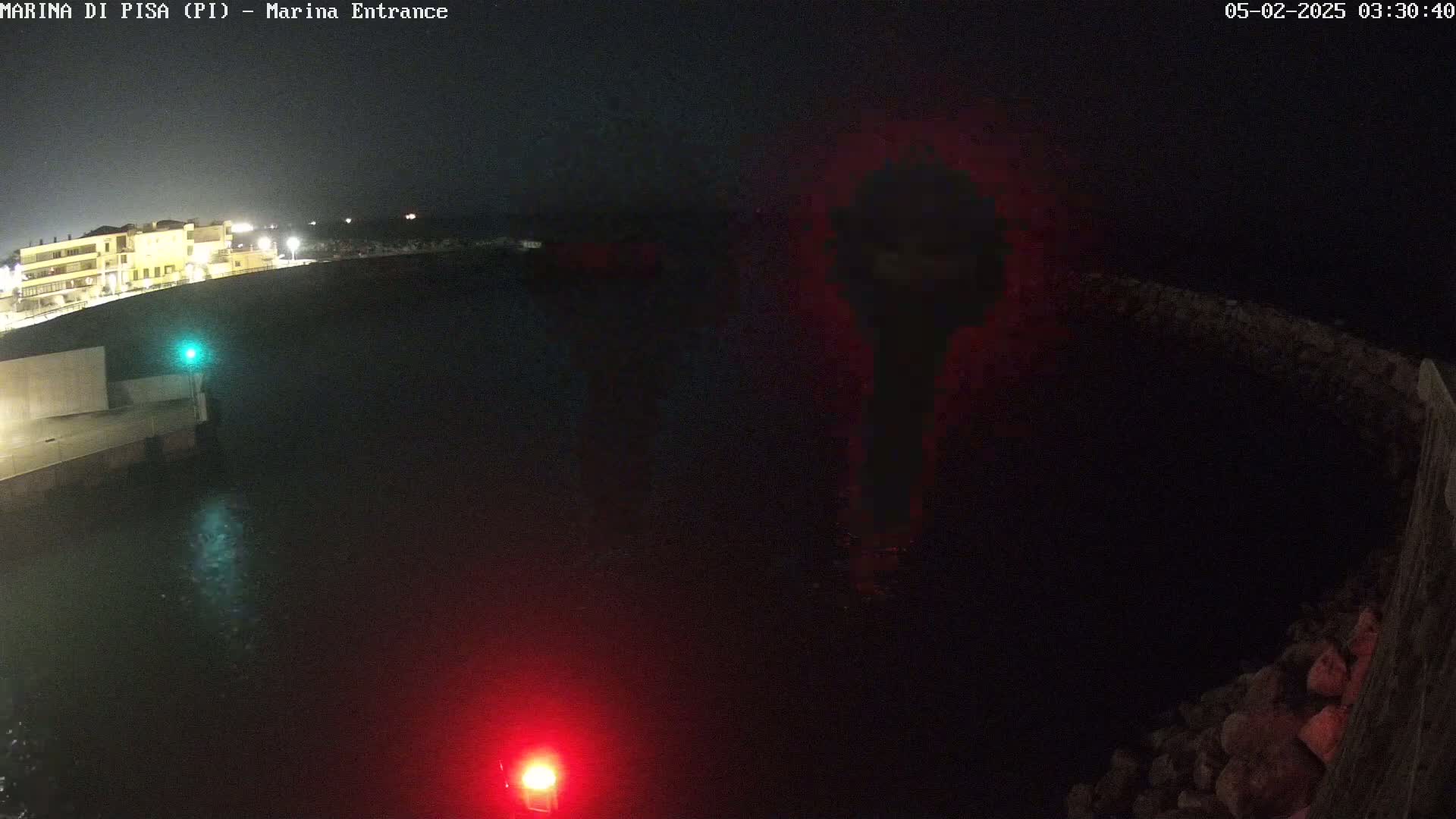 A nighttime view of a dark harbor with a building illuminated on the left, a red and a green light visible on the water, and dark stone walls bordering the water.