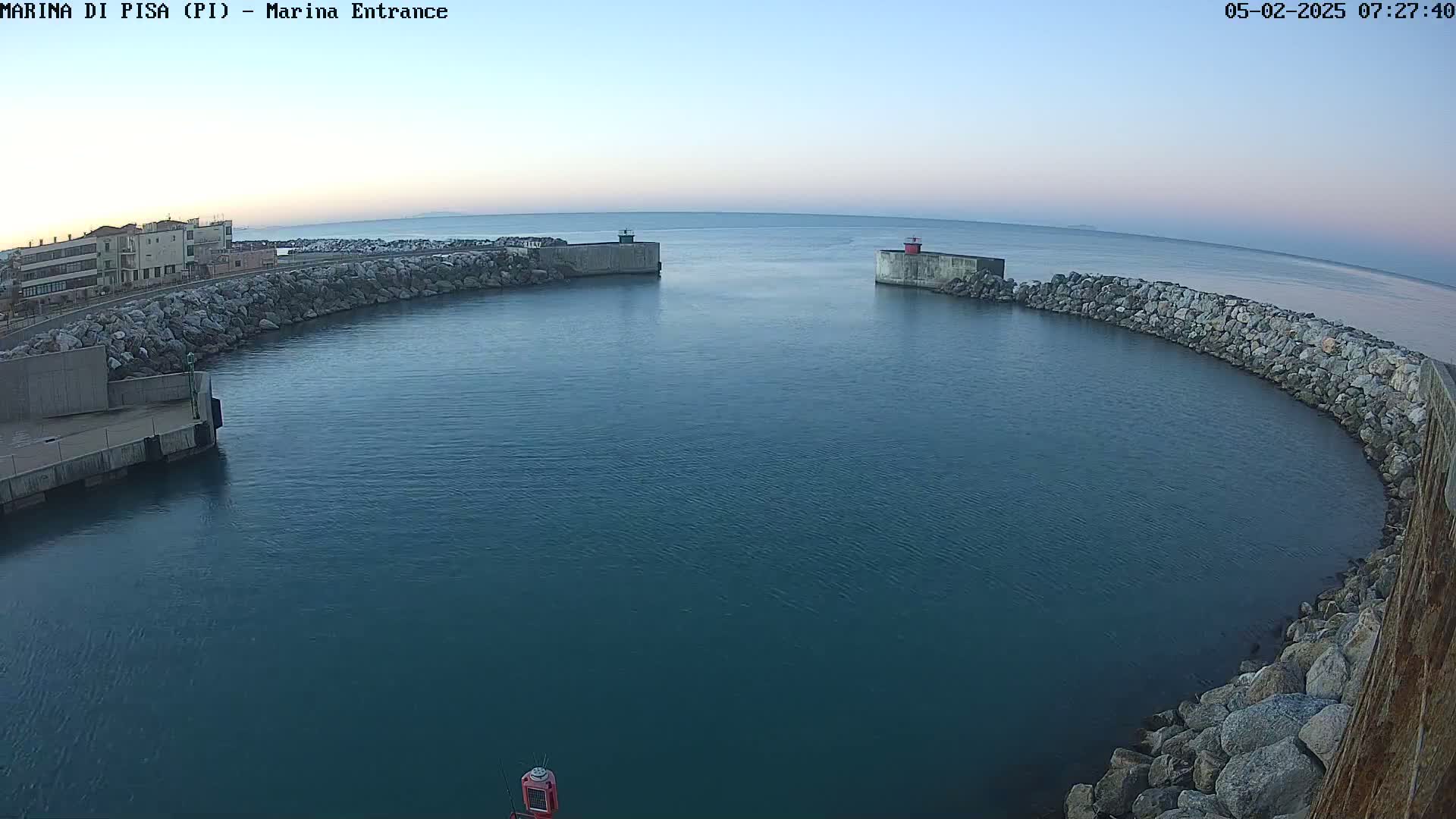 A calm, blue harbor is enclosed by a curved rock wall with a building visible on the left under a pale blue sky.