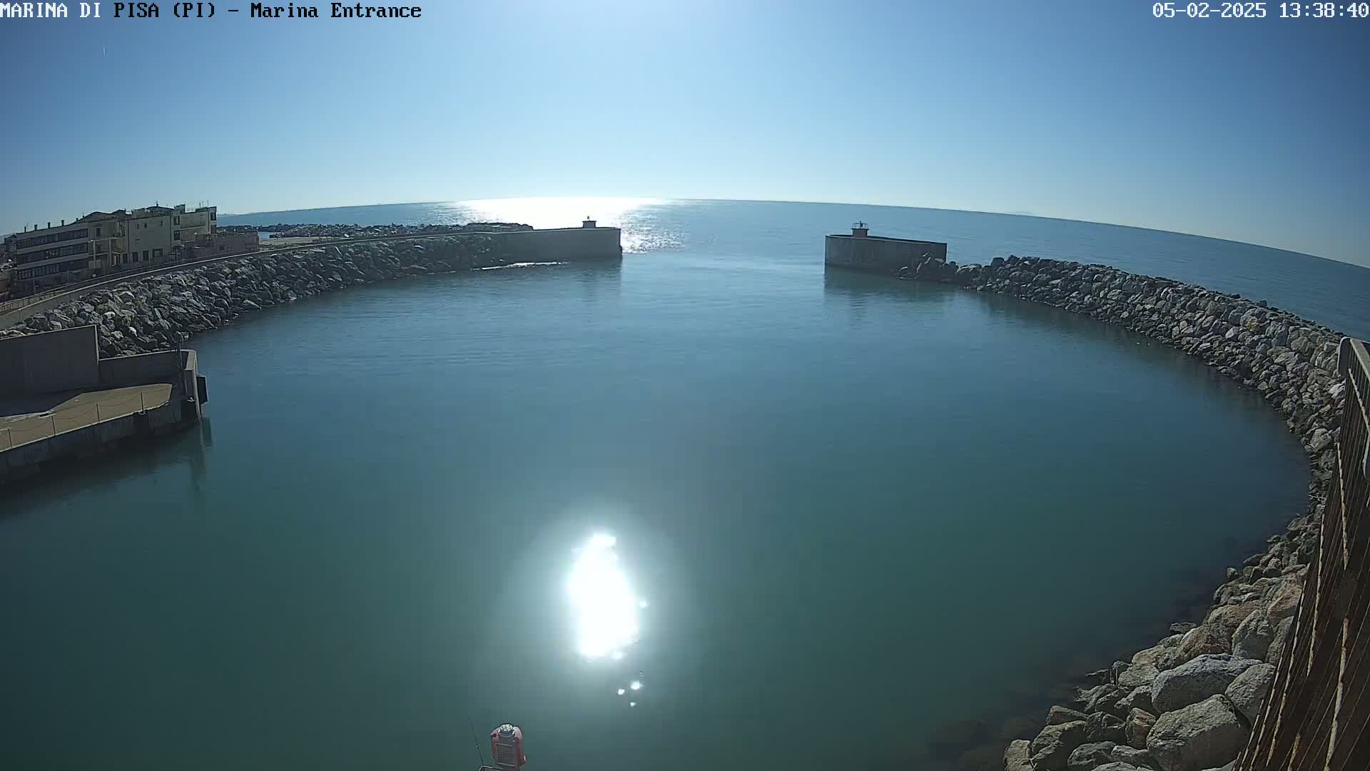 Under a clear, sunny sky, a calm teal harbor is enclosed by stone walls and breakwaters, with a building visible in the distance.