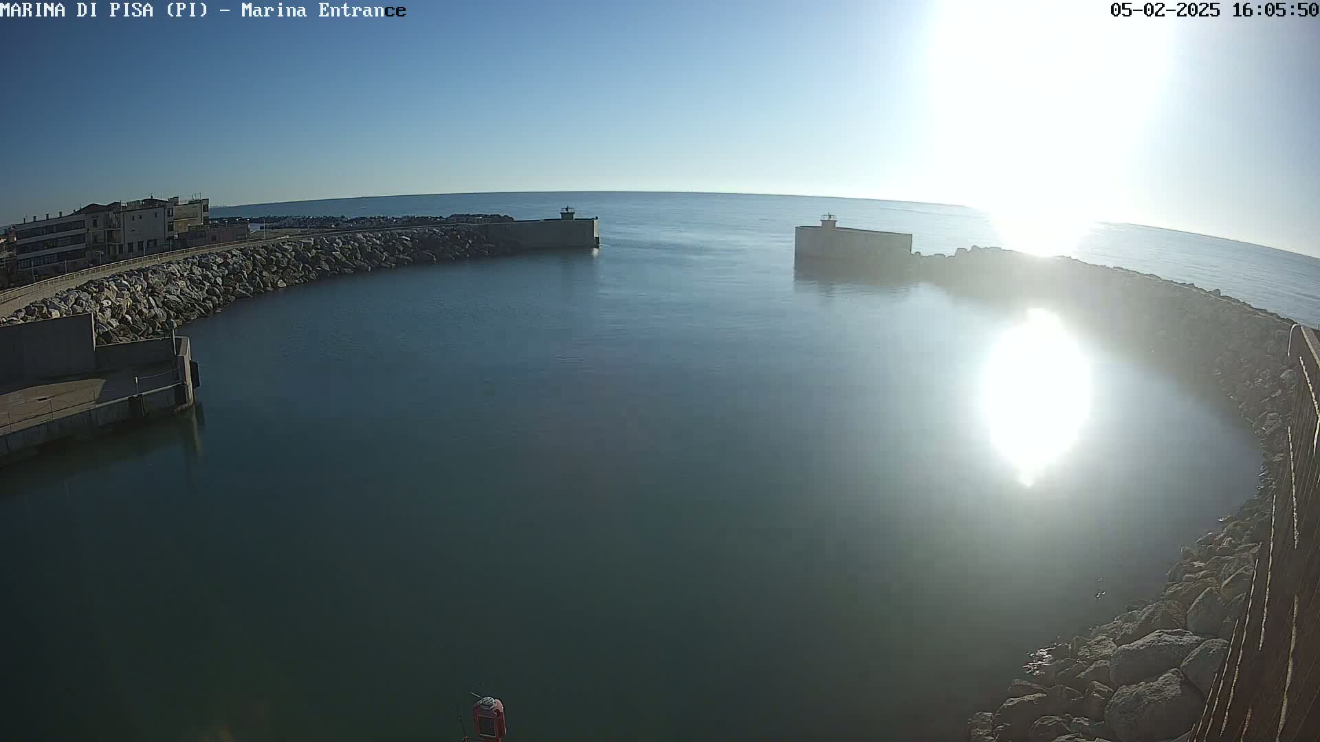 A calm, sunlit harbor is enclosed by rocky breakwaters, with a building visible in the background.