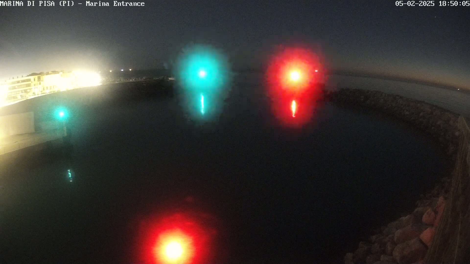 At night, a dark body of water is illuminated by three bright lights—two red and one green—reflected on its surface near a stone wall and a building.