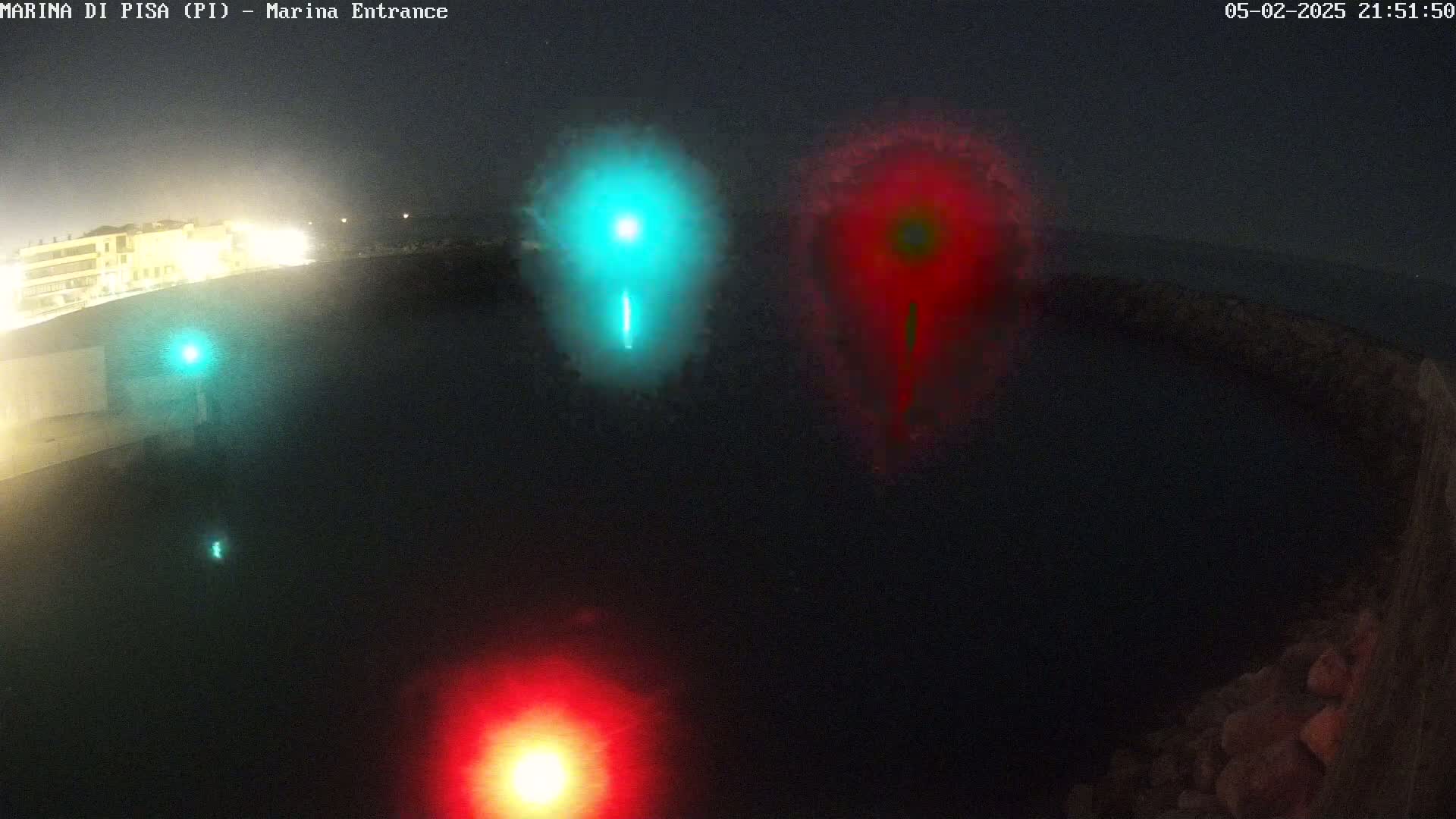At night, three blurry lights, one teal and two red, illuminate a dark body of water near a dimly lit shoreline.
