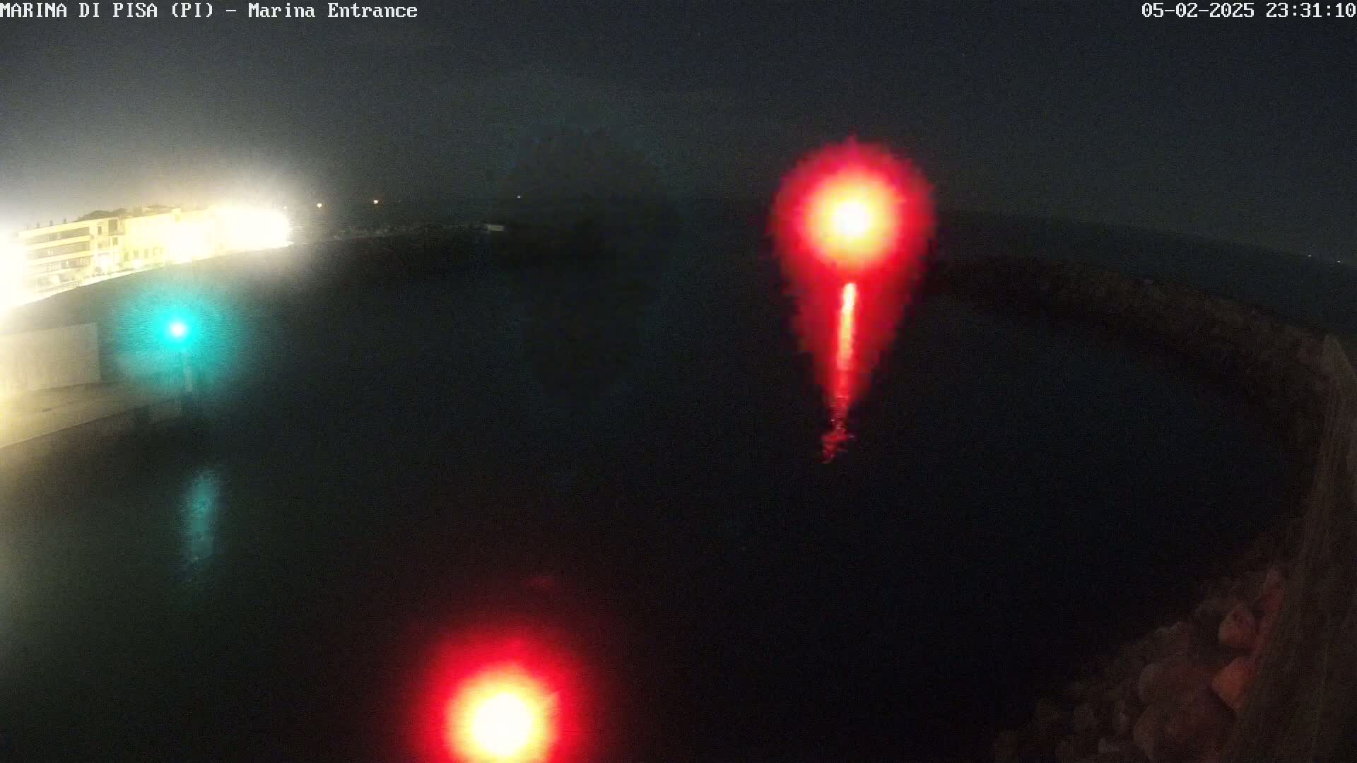 At night, two bright red lights and a smaller green light are visible on dark water near a shoreline.