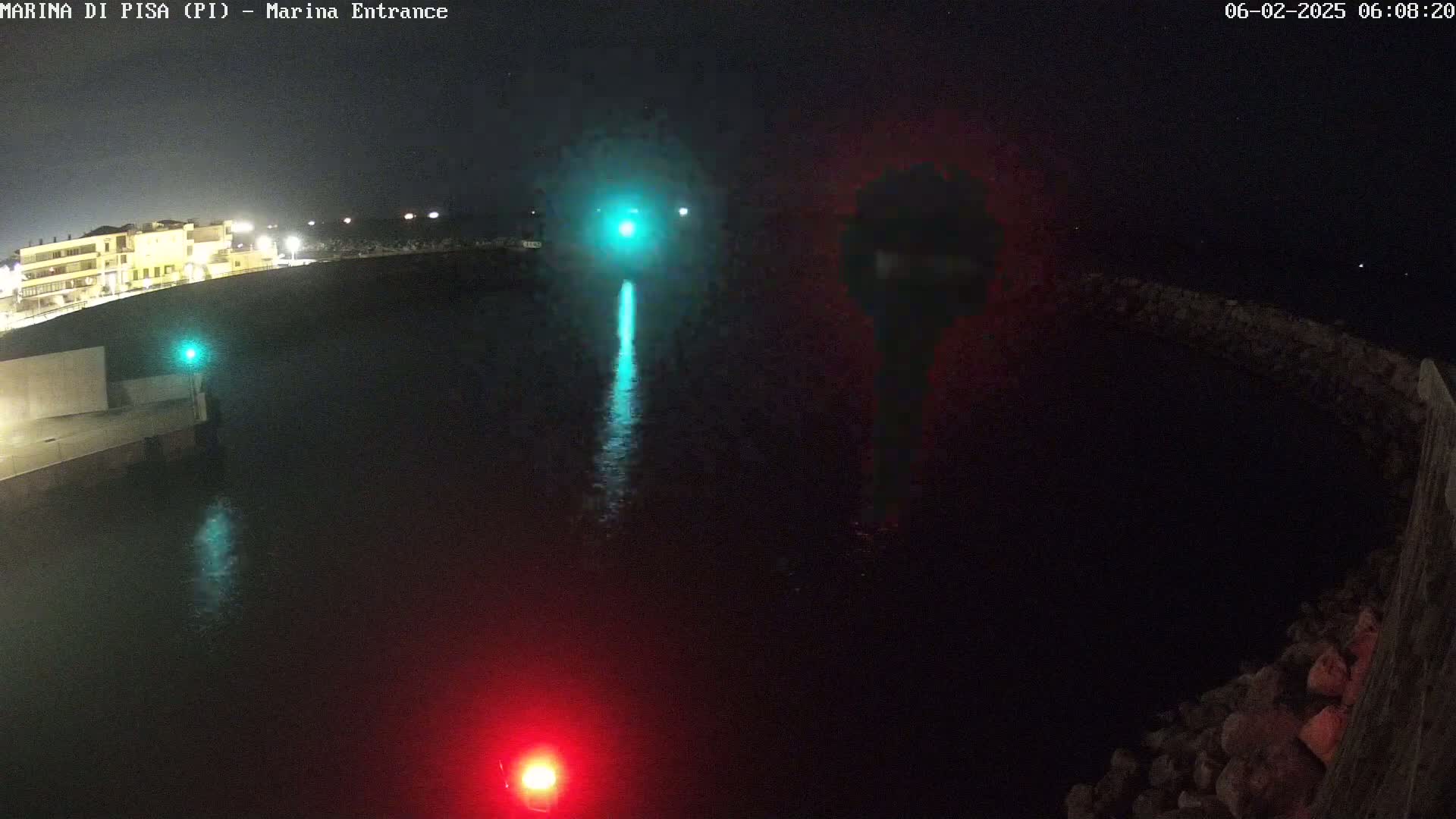 A nighttime aerial view of a dark harbor with a green and red light, and a building illuminated in the distance.