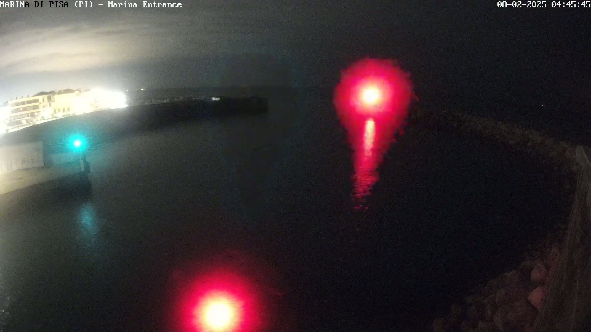 A nighttime view of a marina entrance shows dark water with two red lights and a green light reflecting on the surface.