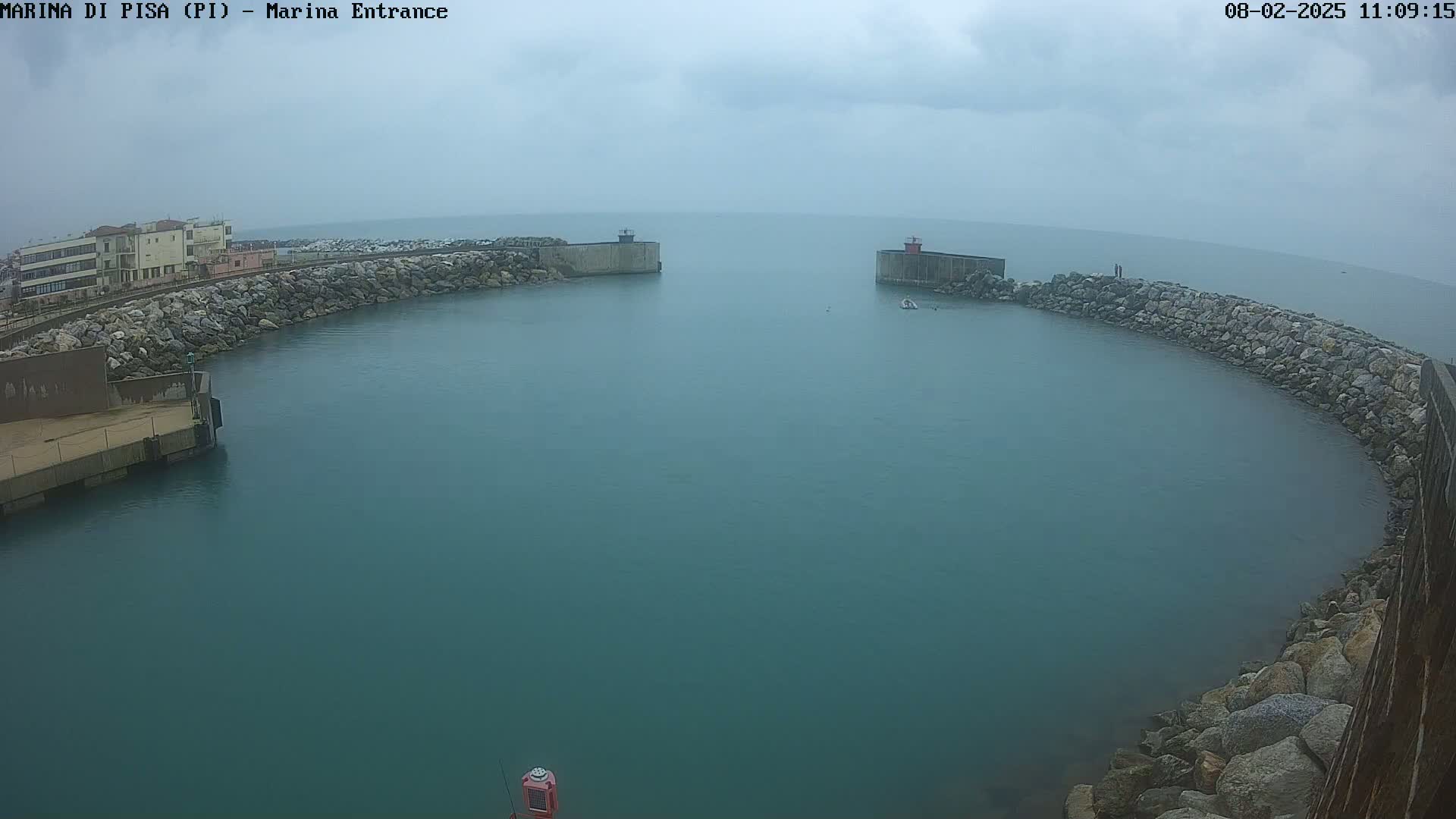 A calm, overcast day shows a harbor entrance with stone walls and buildings in the distance.