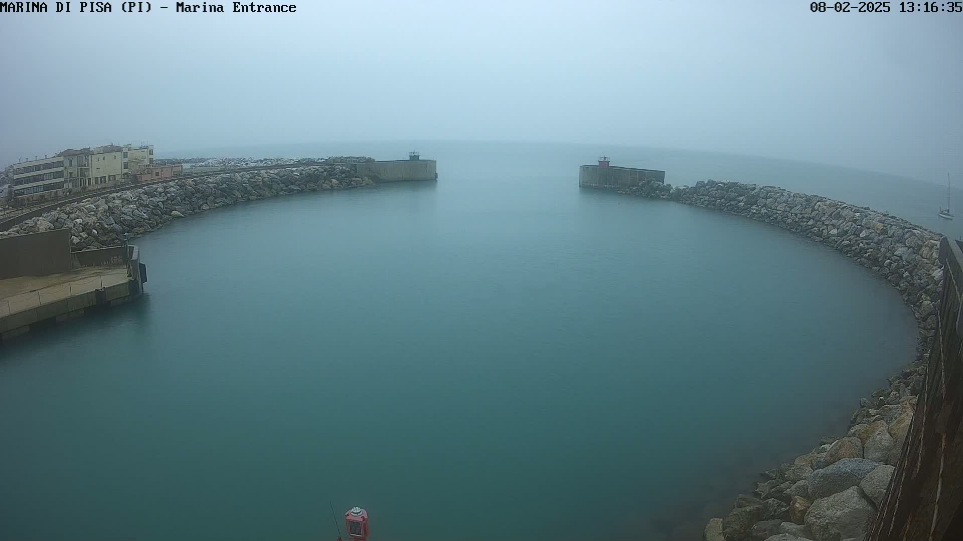 A harbor entrance with calm, teal water is partially shrouded in fog, bordered by rock walls and a building in the distance.