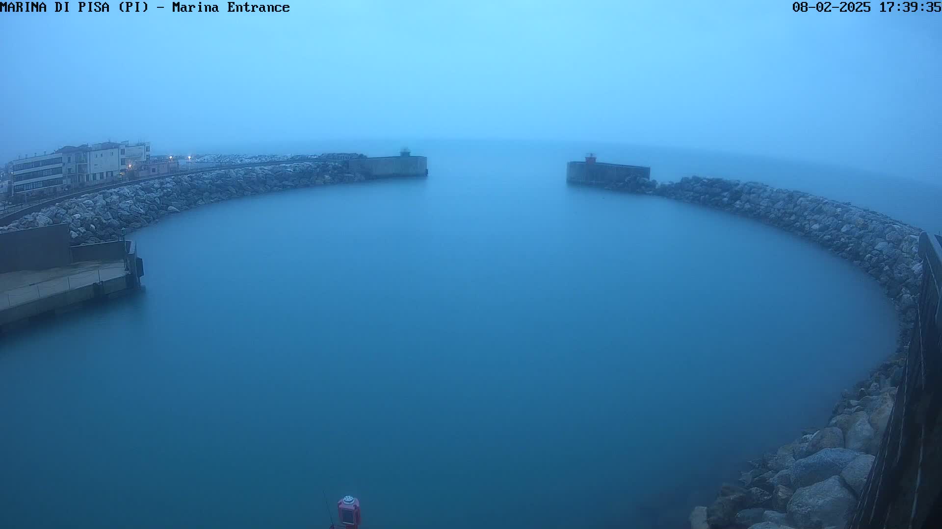 A calm, hazy body of water is enclosed by a curved rock wall, with buildings visible in the distance under a misty sky.