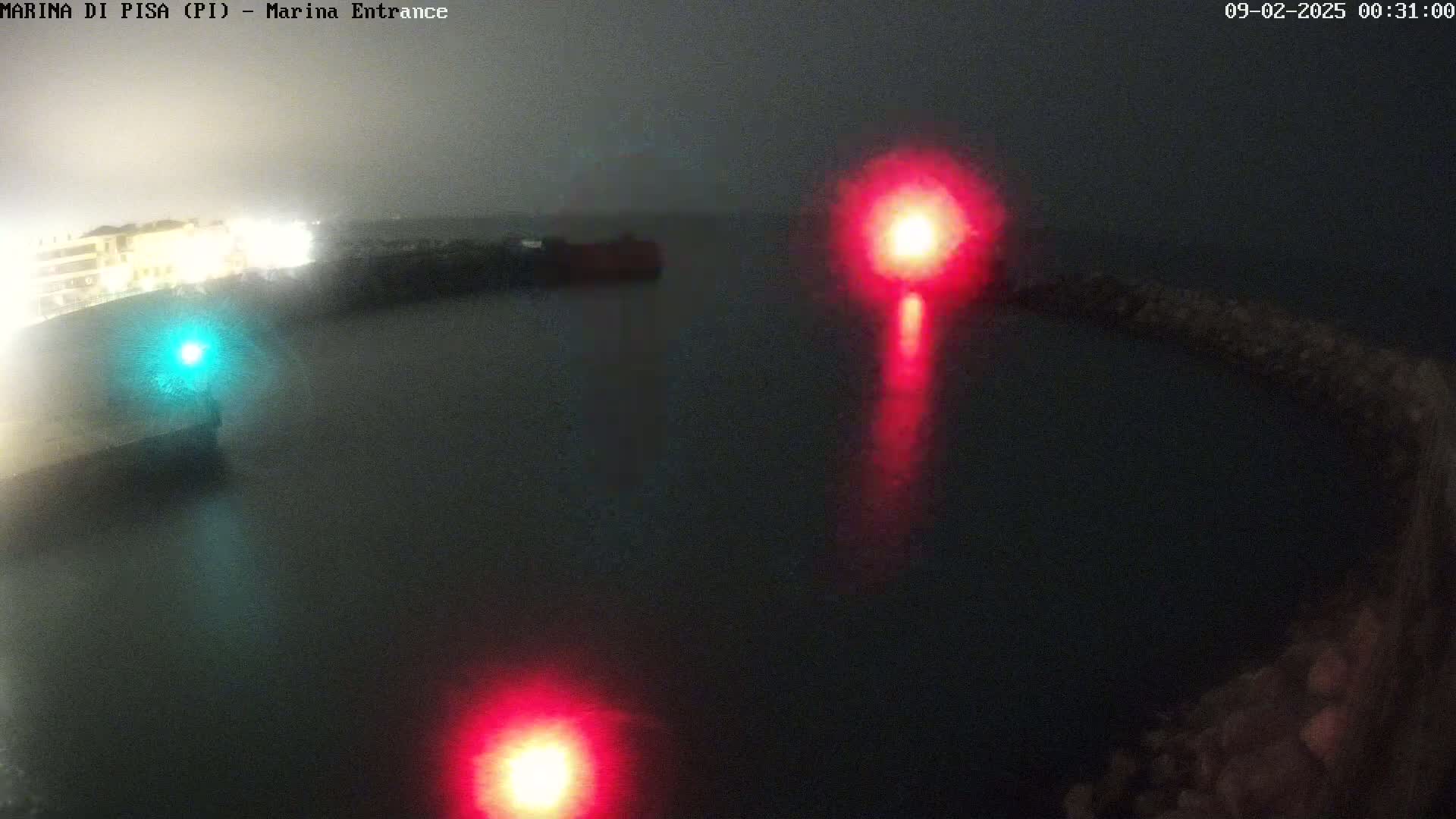 A nighttime, foggy view of a harbor entrance with red and green navigation lights visible on the water.