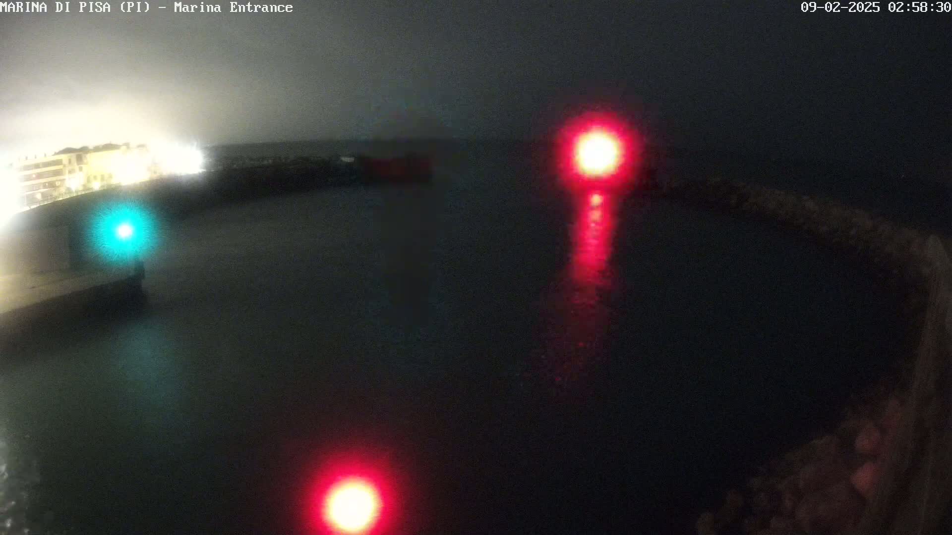 At night, a dark body of water is illuminated by a green light on the left and two red lights on the right, with a blurry building visible in the background under a hazy sky.