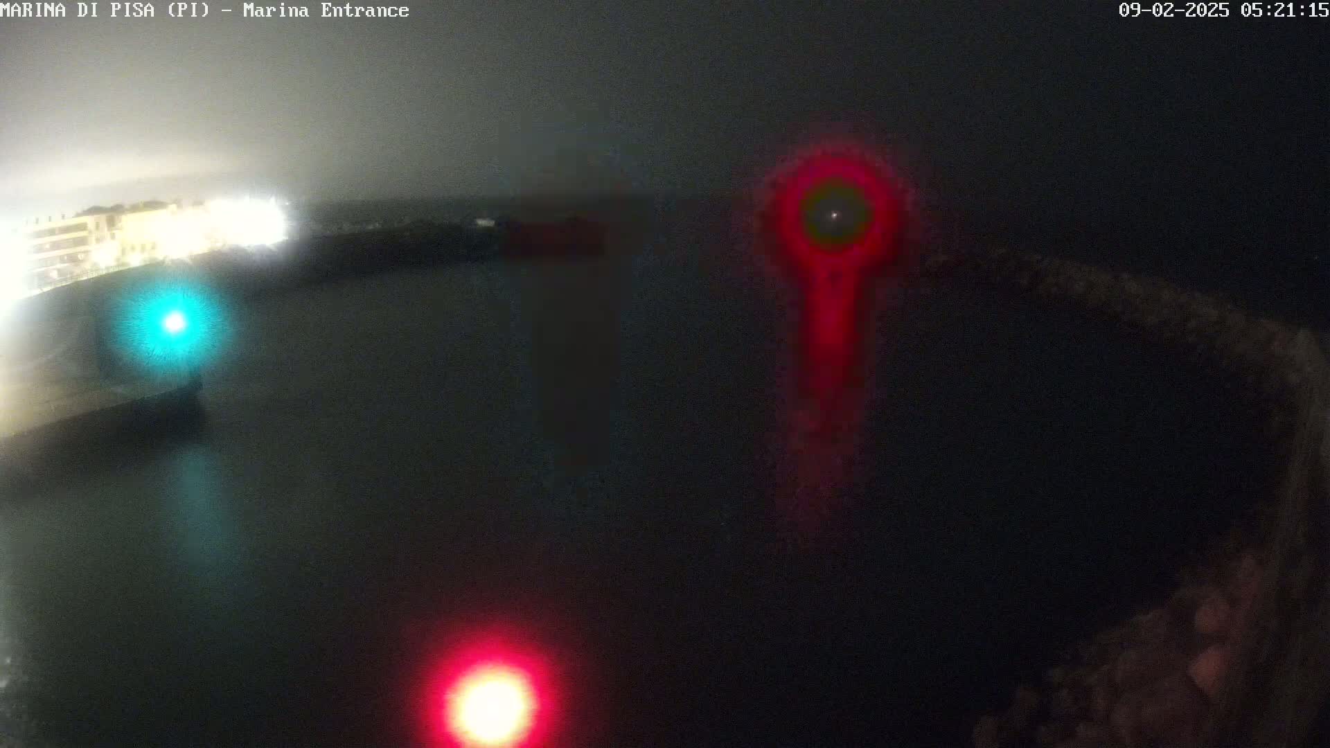 A nighttime, dark view of a harbor entrance with a bright red light and a fainter green light visible in the water.