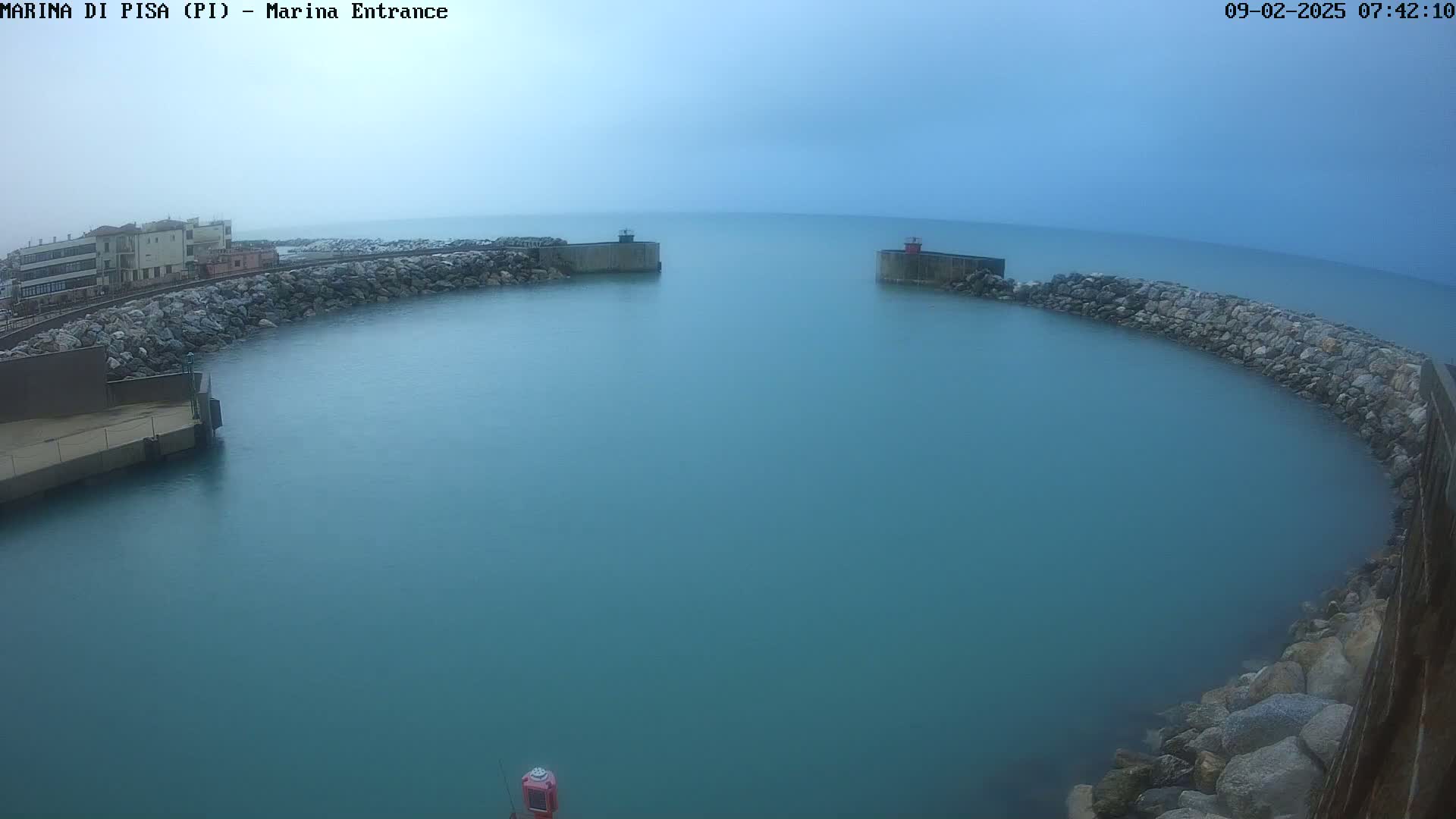 A calm, teal harbor is partially enclosed by rock walls and a building is visible in the distance under an overcast sky.