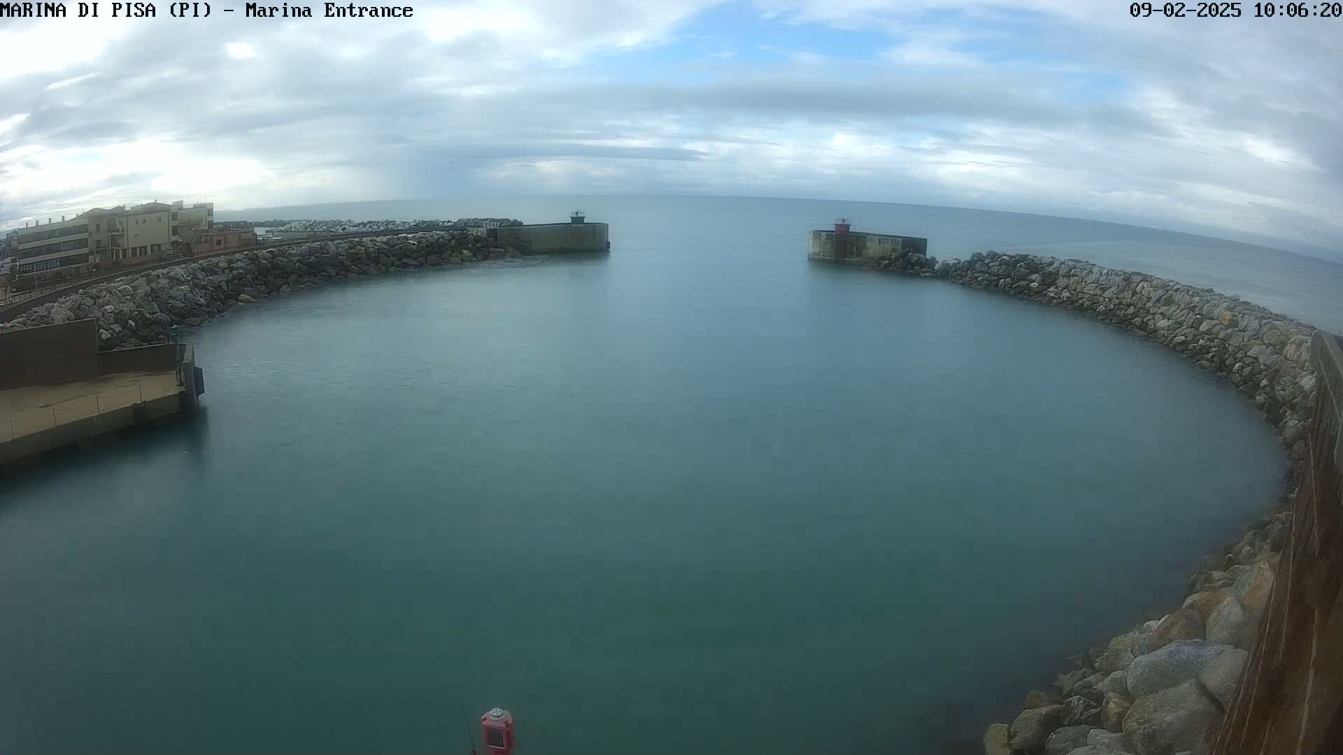 A calm, partly cloudy day shows a harbor entrance with stone walls and buildings on one side, and a small red structure on the other.