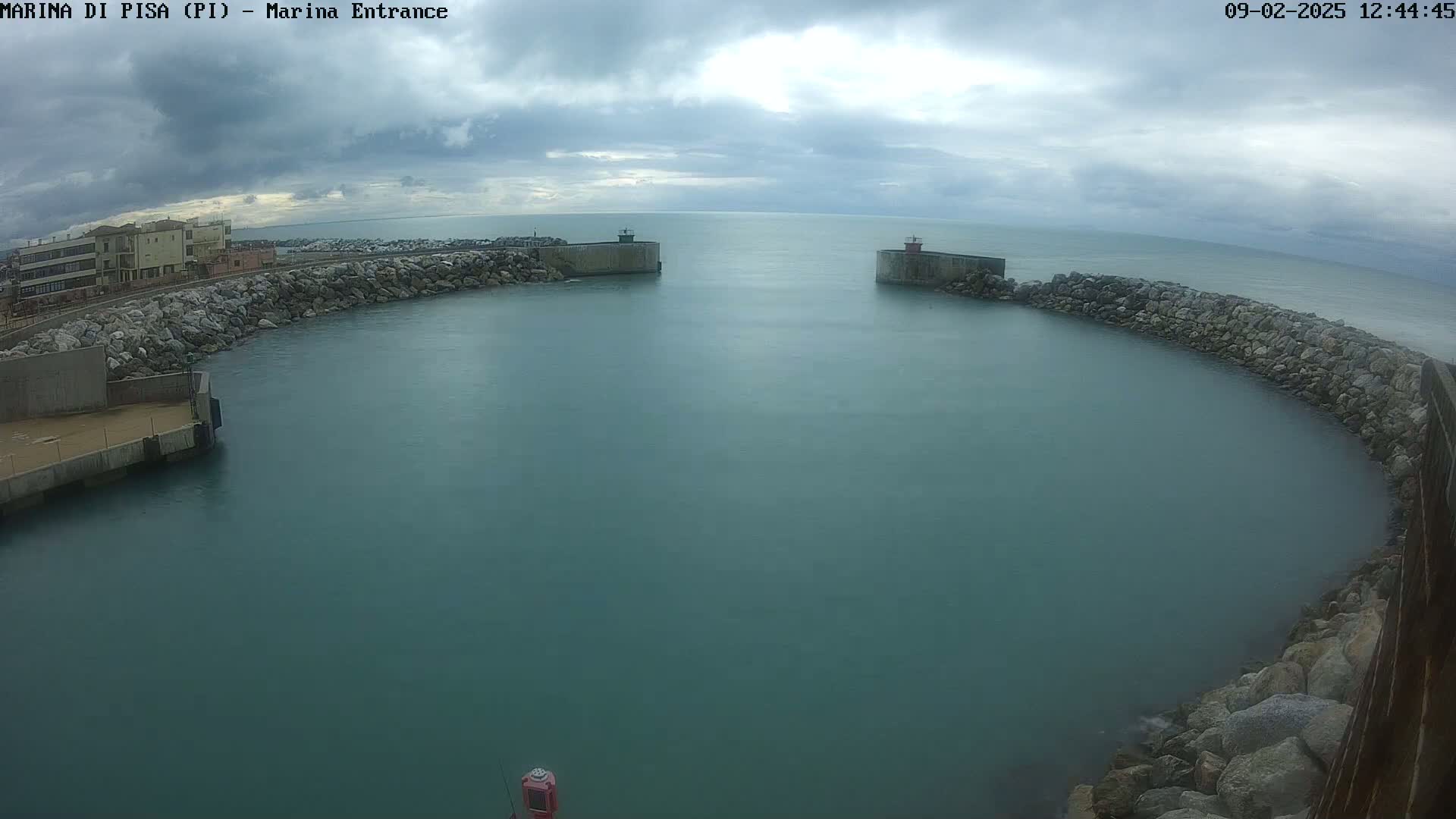 A calm, overcast day reveals a harbor entrance with stone walls and buildings in the background.
