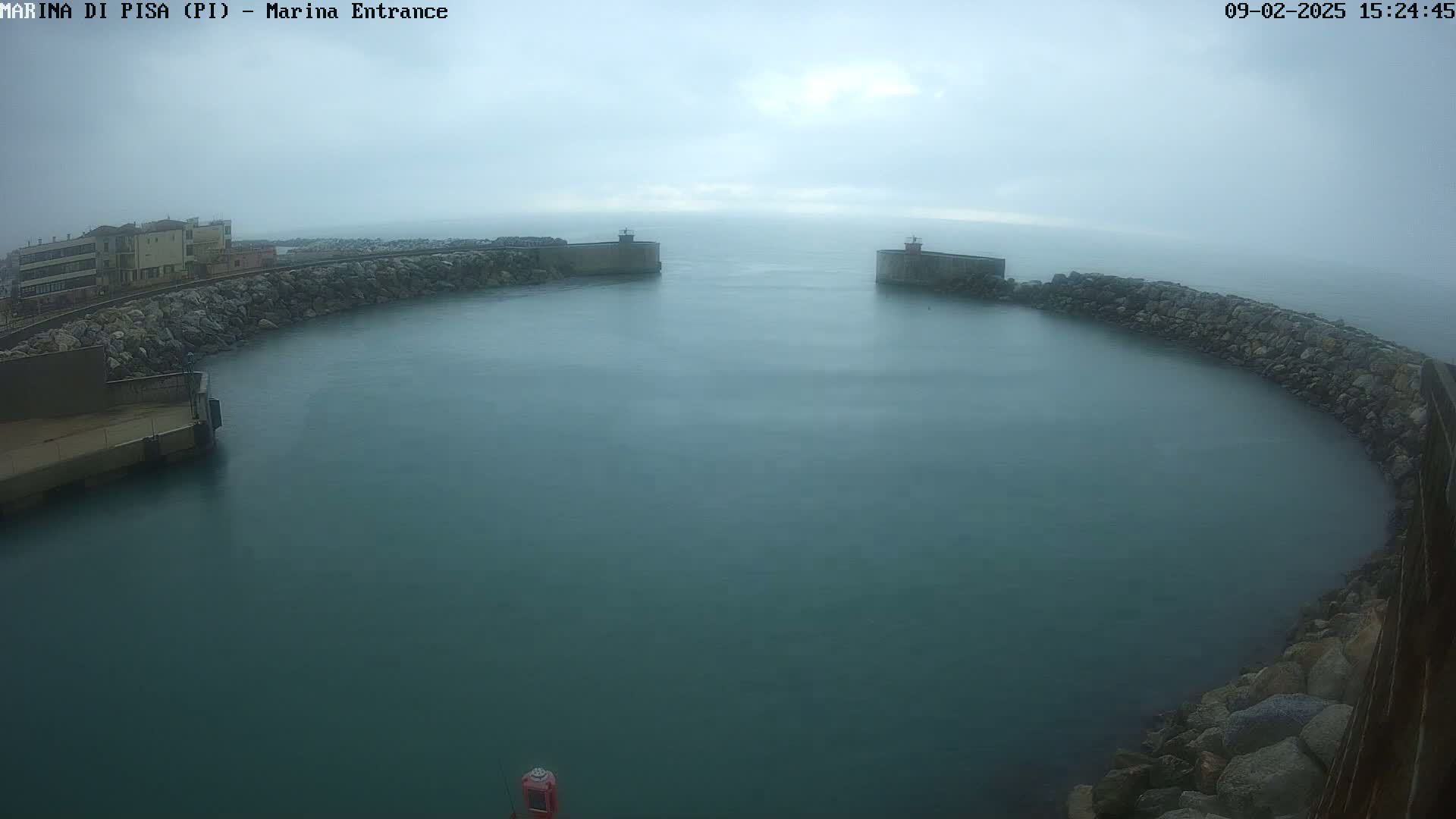 A harbor entrance with calm, murky water under an overcast sky is seen from an elevated vantage point.