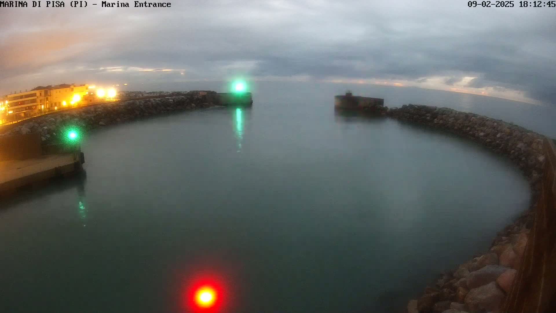 A harbor entrance at twilight under a cloudy sky, showing calm water enclosed by rock walls and illuminated by lights on shore and in the water.
