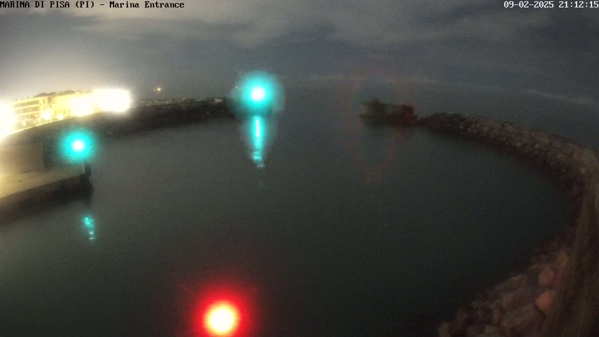 A nighttime view of a harbor entrance shows calm dark water with a few lights reflecting on the surface, and a stone breakwater curving into the scene.