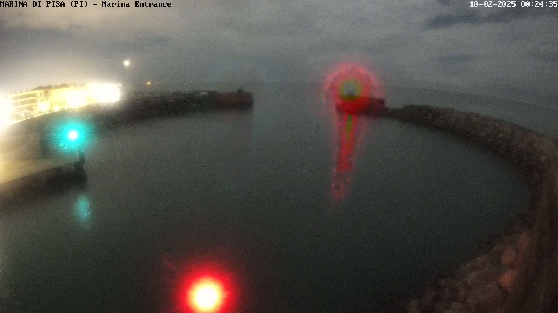 A nighttime view of a harbor entrance shows calm, dark water with lights reflecting on the surface, illuminated structures along the shore, and a blurry, multicolored light source in the middle distance under an overcast sky.